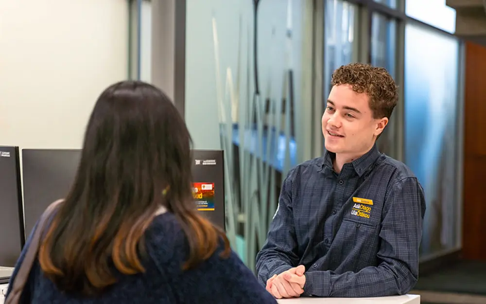 A staff member at the AskOtago help desk smiles while assisting a student.