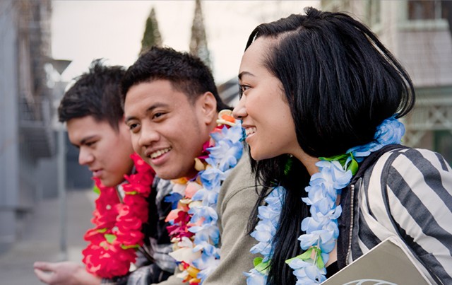 Three students wearing bright flower leis smile while sitting together outdoors.