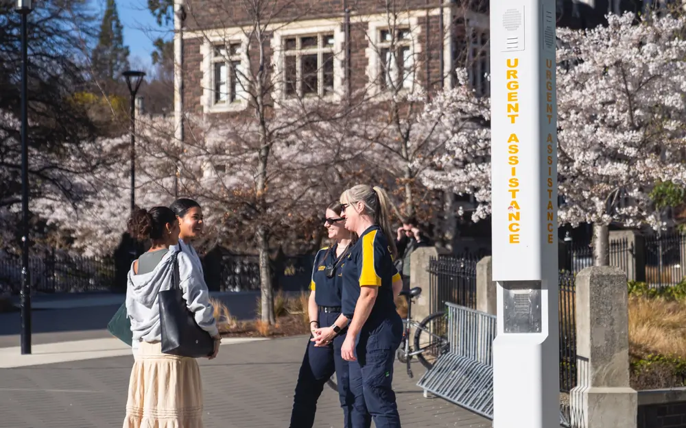 Campus Watch staff in uniform talk with students on campus next to an emergency assistance pillar.