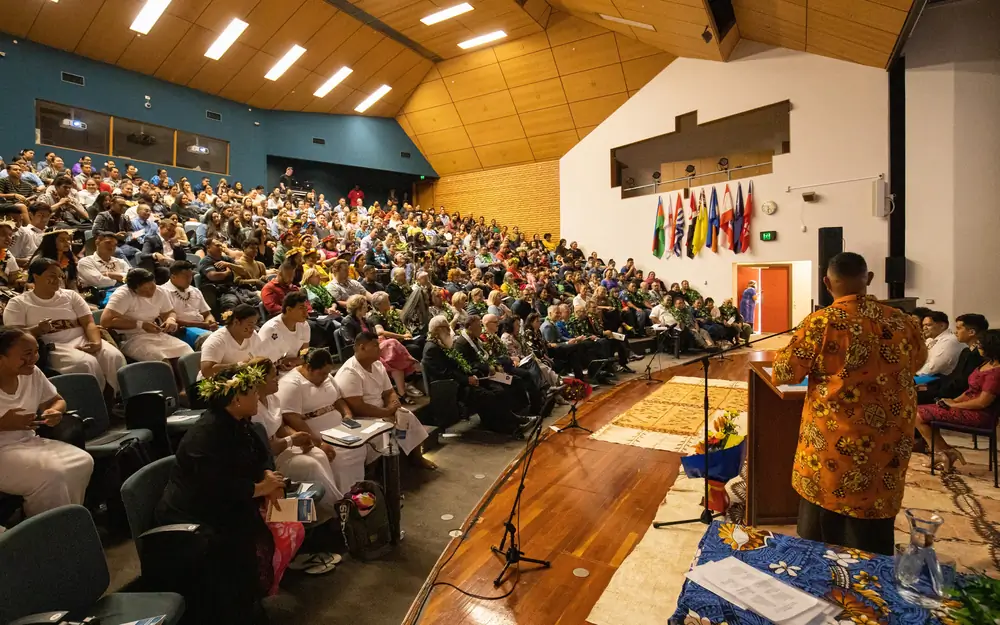 A speaker addresses a large audience in a lecture hall decorated with Pacific nation flags.