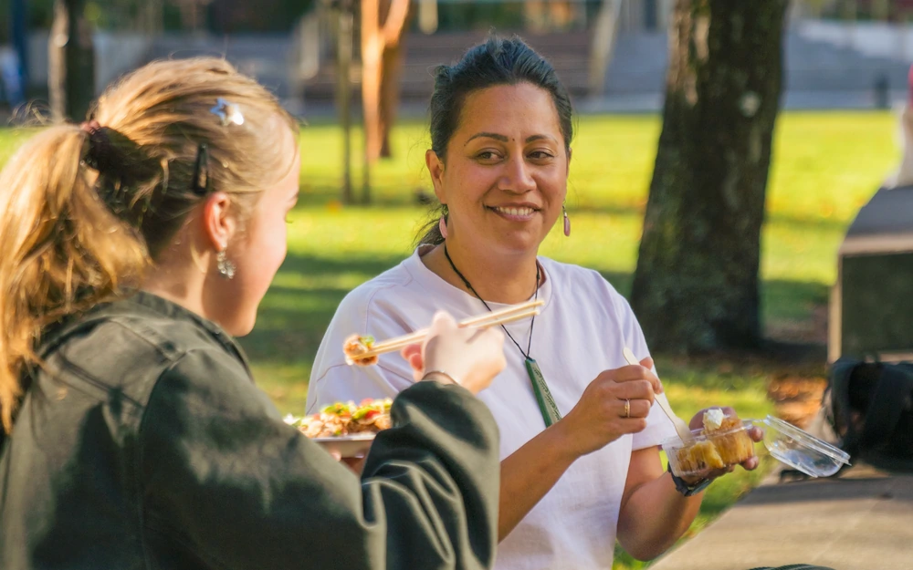 Two students share a meal outdoors in a sunny park setting.