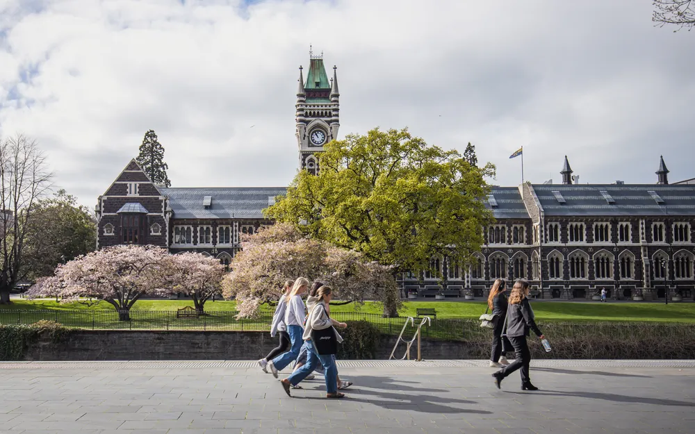 Students walk past the historic University of Otago Clocktower building during spring, with blossoming cherry trees in the foreground.