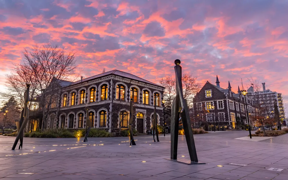 The historic University of Otago Staff Club building stands beside a modern sculpture under a vibrant pink and orange sunset.