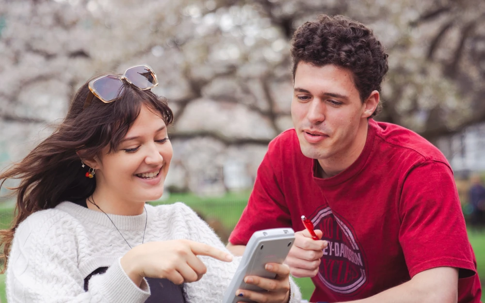 Two students smile while looking at a graphing calculator screen outdoors under cherry blossoms.