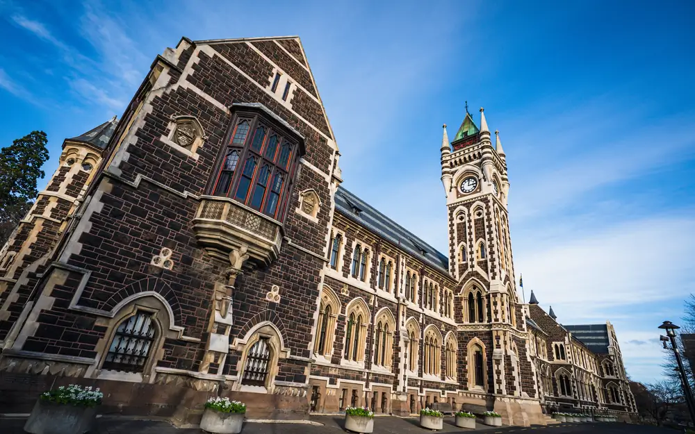 A wide angle view of the University of Otago Clocktower building featuring dark stone walls and a white clock tower against a clear blue sky.