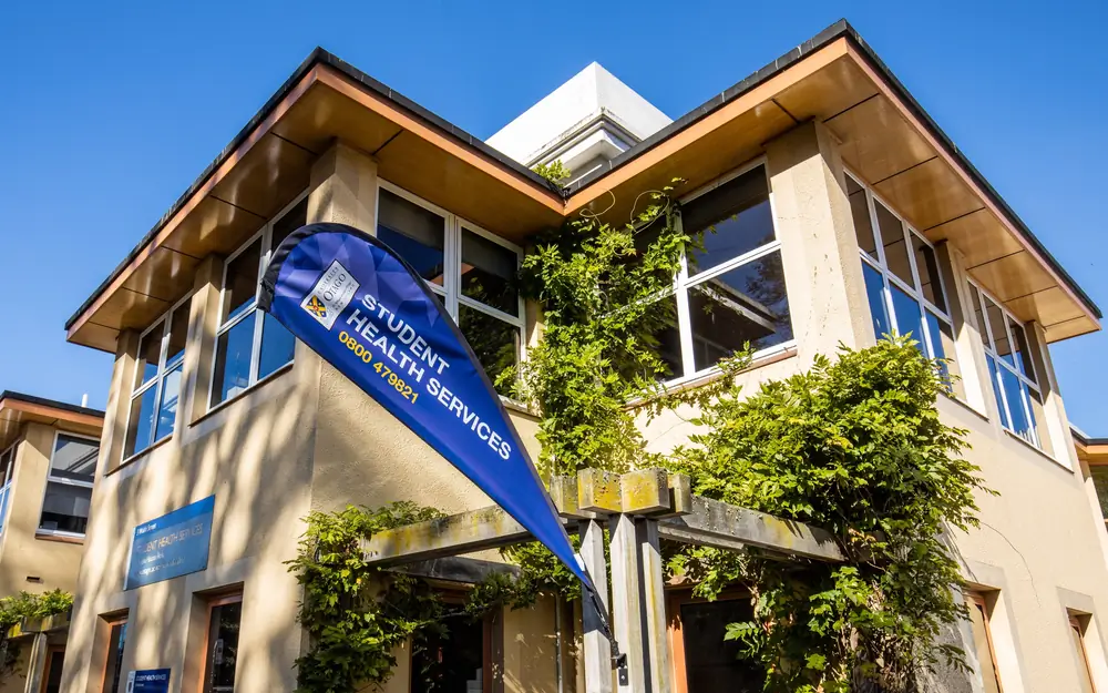 The University of Otago Student Health Services building with a blue flag in the foreground under a clear sky.