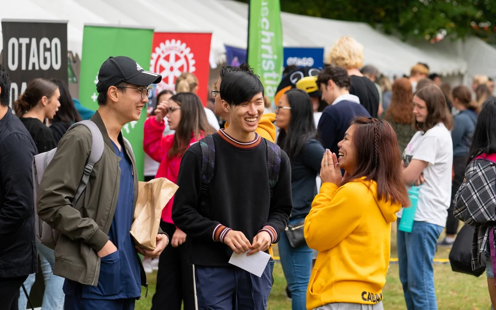 Three students laugh and talk at a crowded outdoor university club fair with colorful banners and tents in the background.