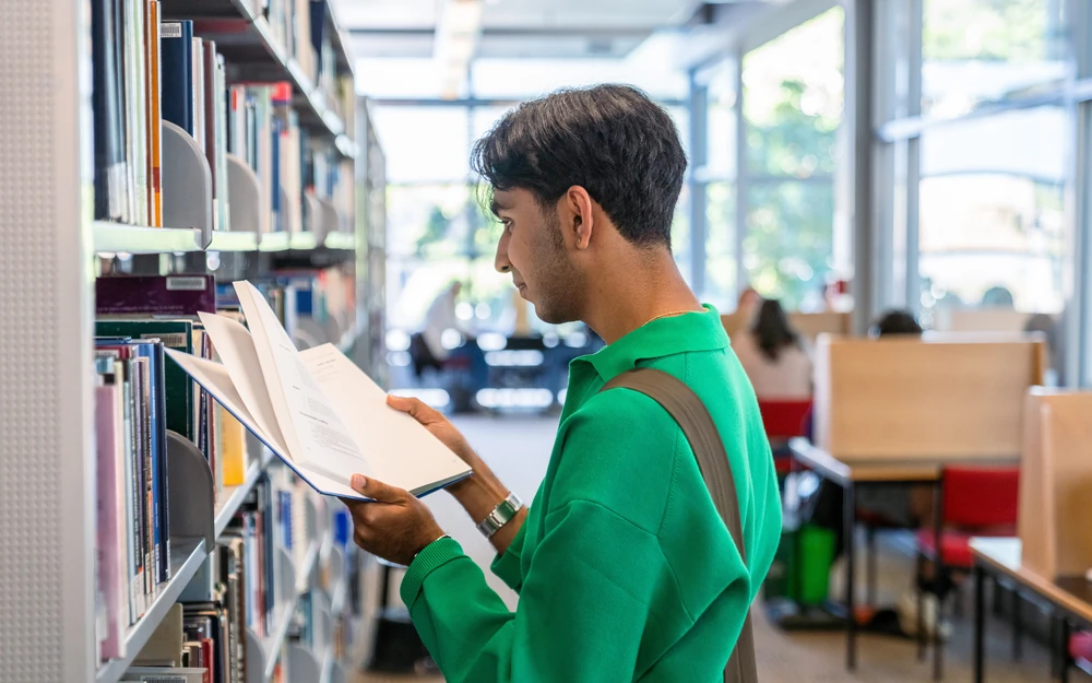 A student in a green shirt browses a book while standing between shelves in a university library.