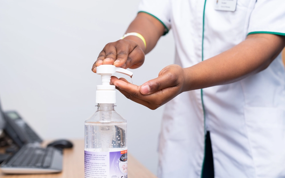 A person wearing a white uniform with green trim presses the pump of a hand sanitizer bottle to dispense gel into their palm.