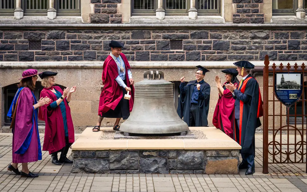 Academics in graduation regalia gather around a large bronze bell as one individual strikes it with a mallet.