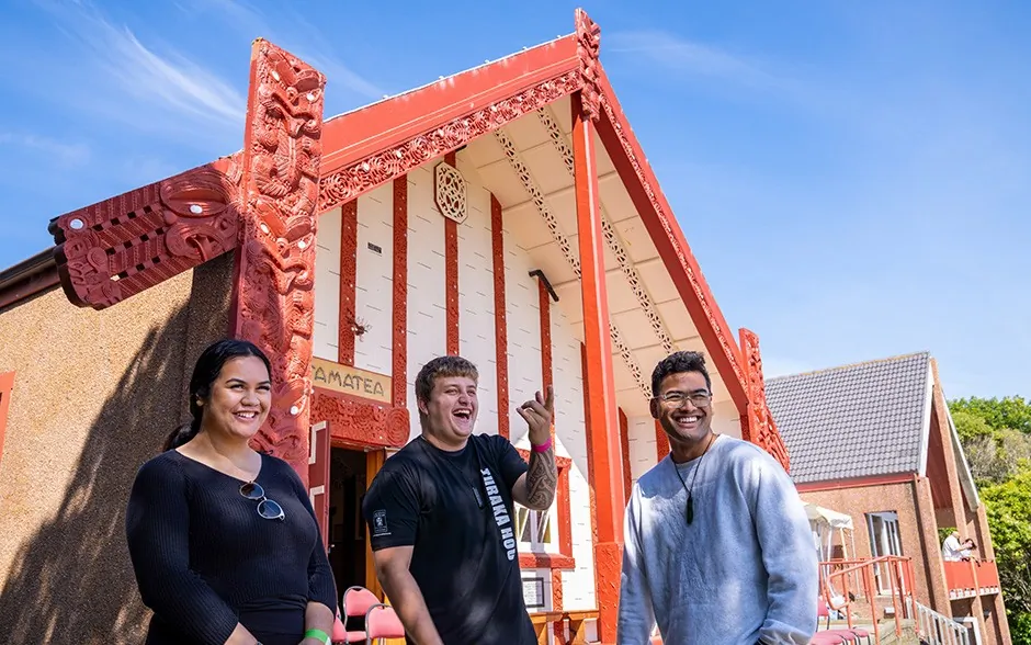 Three students smile in front of the intricately carved meeting house at Ōtākou Marae.