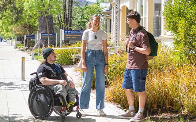 Three students chat on a sunny campus sidewalk with one student using a wheelchair.