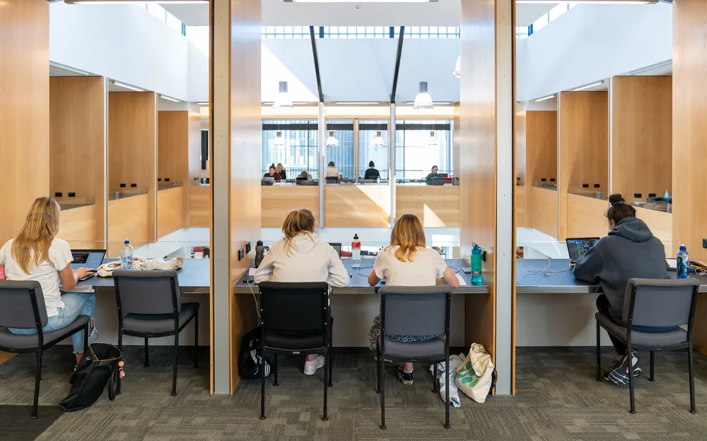 Students study at modern wooden desks in a bright, multi level university library.