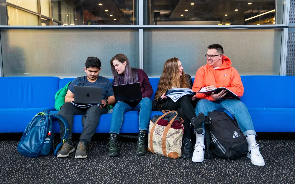 Four students sit together on a long blue couch while working on laptops and reviewing notes.