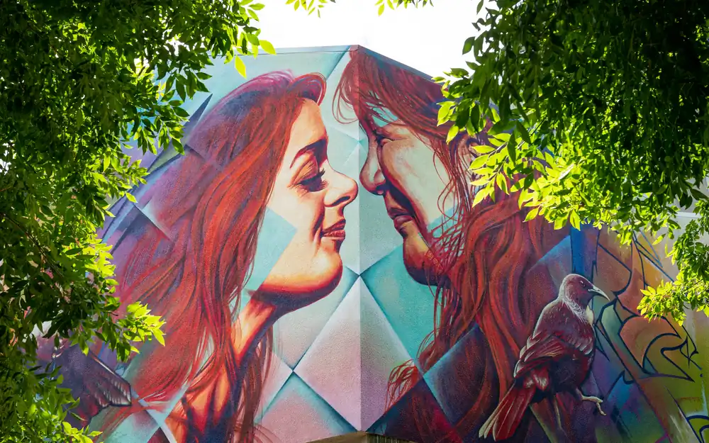 A large mural on a building corner shows the faces of two women looking at each other, framed by green tree branches.