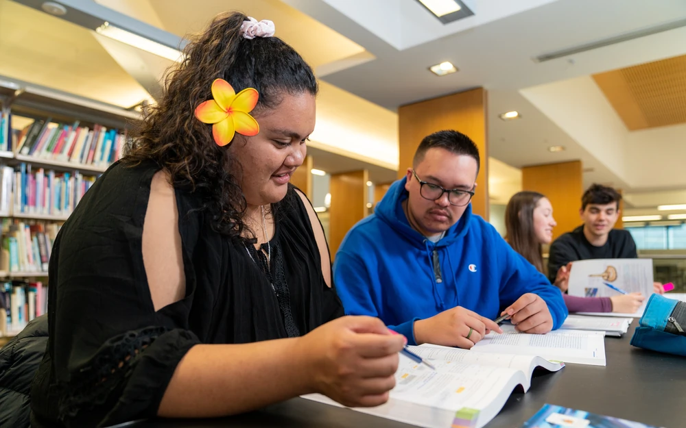A student with a frangipani flower in her hair studies a textbook at a library table with other students.