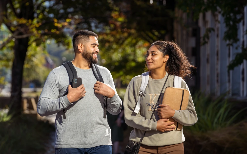 Two students with backpacks walk and talk together in the sunlight.