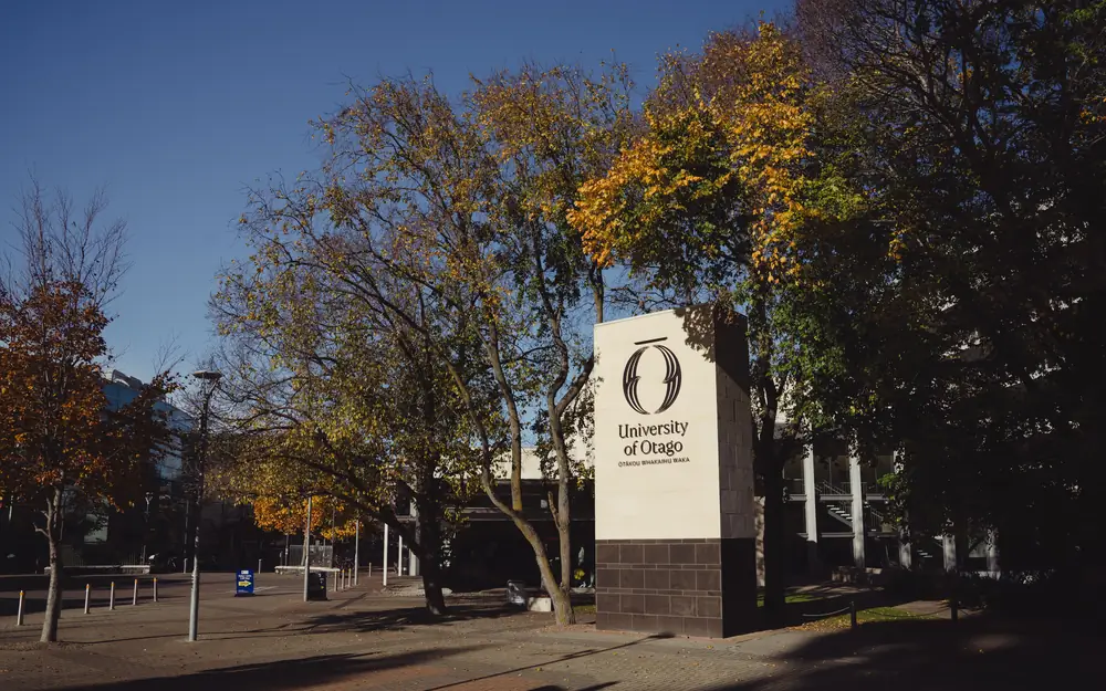 A University of Otago entrance sign made of white stone stands among trees with autumn leaves.