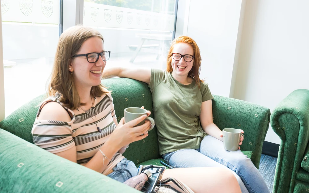 Two students laugh and share a conversation while holding mugs on a green sofa.
