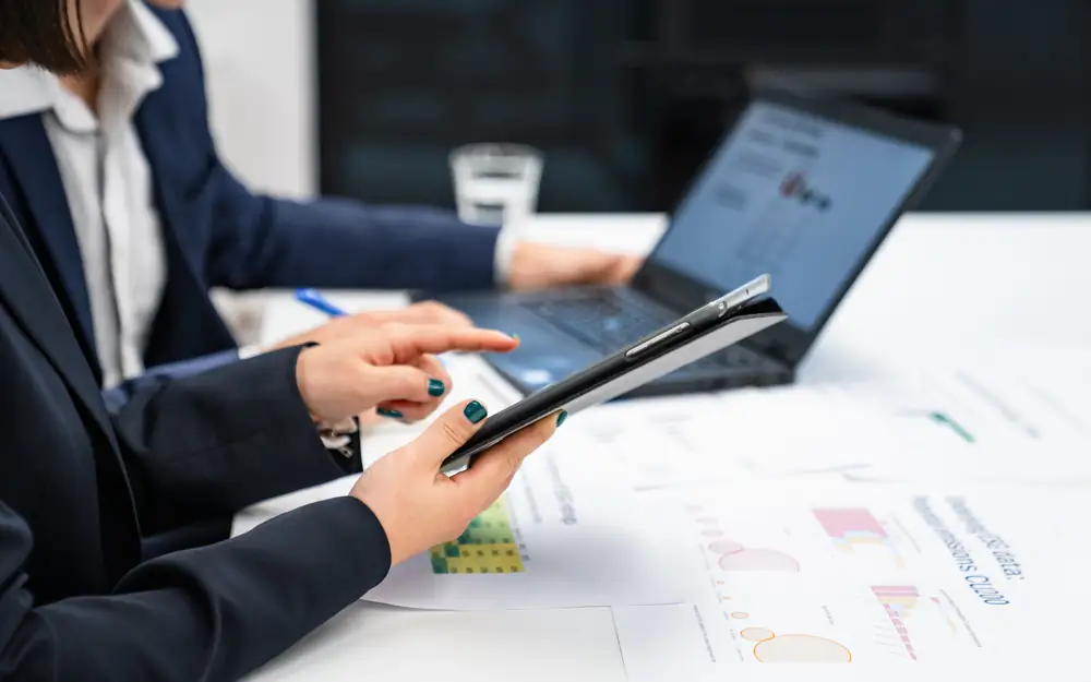 A person uses a tablet at a desk while working alongside a colleague with a laptop.