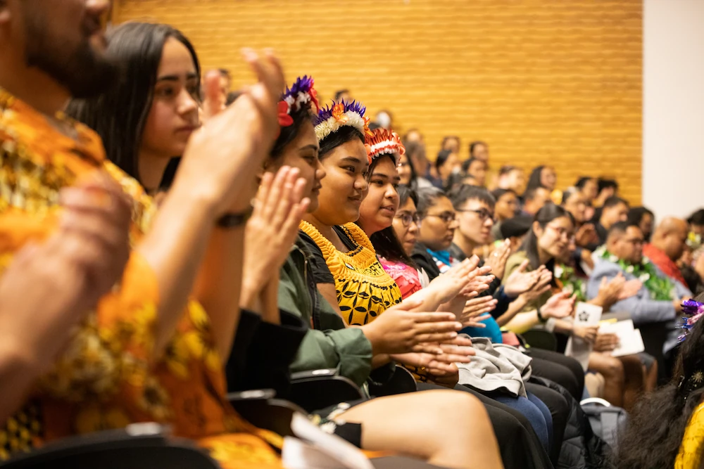 A side view of a diverse audience in an auditorium. Several students in the foreground wear traditional Pacific Island floral headpieces and patterned clothing while smiling and clapping.