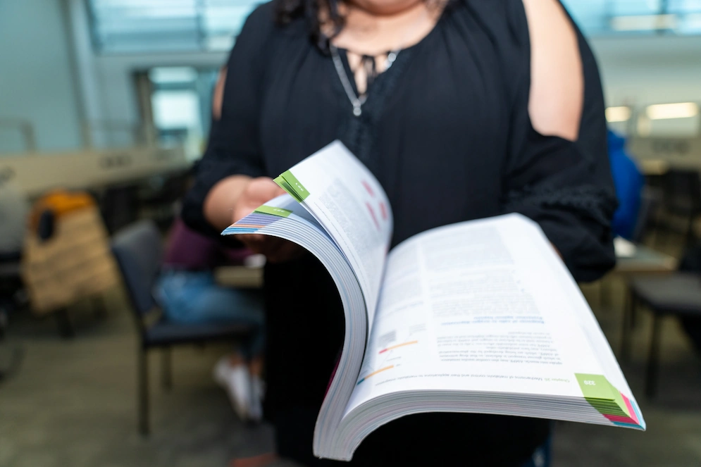 A person in a black top flips through a thick, open textbook with colorful page tabs. The background shows a blurred library or study hall setting.