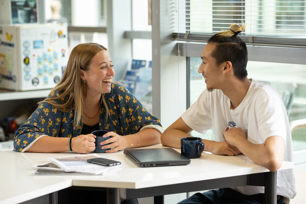 Two students sit at a white table with mugs and a laptop, engaged in a cheerful conversation. One woman laughs while a man with a top knot smiles back.